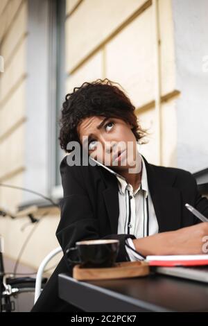 Portrait eines afroamerikanischen Mädchens, das am Tisch des Cafés sitzt und auf ihrem Handy spricht, während es ertrampt zur Seite schaut. Nachdenkliche Dame mit dunklem Cur Stockfoto
