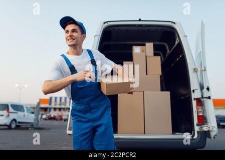 Deliveryman in Uniform hält Pakete am Auto, Lieferservice, Lieferung. Mann steht an Pappverpackungen im Fahrzeug, männlich liefern, Kurier oder Stockfoto
