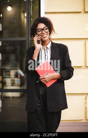 Lächelndes afroamerikanisches Mädchen in Gläsern, das neben der Tür steht und glücklich beiseite schaut, während es auf ihrem Handy spricht. Lady mit dunklen lockigen Haaren in Sui Stockfoto