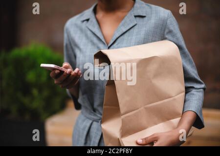 Nahaufnahme Foto von Frau Körper in grauem Kleid stehen und halten Papierbeutel. Foto von Frau Hände mit Handy und Papiertüte auf der Straße Stockfoto