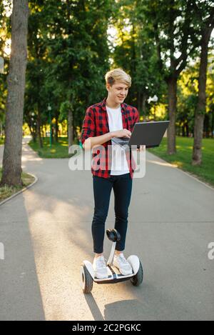 Junger Mann, der auf Mini-Gyro-Board mit Laptop, Sommerpark reitet. Erholung im Freien mit elektrischem gyroboard. Eco Transport mit Balance-Technologie, elect Stockfoto