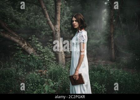 Weibliches Opfer in weißem Kleid hält Buch in der Hand, Wald auf Hintergrund. Foto im Horror-Stil, Geheimnis Stockfoto