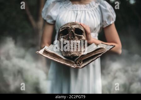 Weibliches Opfer in weißem Kleid hält Buch und menschlichen Schädel in der Hand, Wald auf Hintergrund. Foto im Horrorstil, Exorzismus Stockfoto