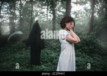 Junge weibliche Opfer in weißem Kleid im Wald, Tod in einem schwarzen Kapuzenpullover und mit einer Sense für den Hintergrund. Foto in Horror Stil, Exorzismus Stockfoto
