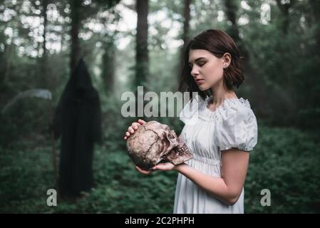 Weibliches Opfer in weißem Kleid hält menschlichen Schädel in den Händen im Wald und Tod. Foto im Horror-Stil Stockfoto