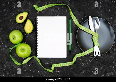 Flache Lage von gekreuzten Gabel und Messer zusammen mit einem Maßband auf einem schwarzen Teller, leeres Papier Notizbuch, Holzstift, frische grüne Äpfel und ein gebunden Stockfoto
