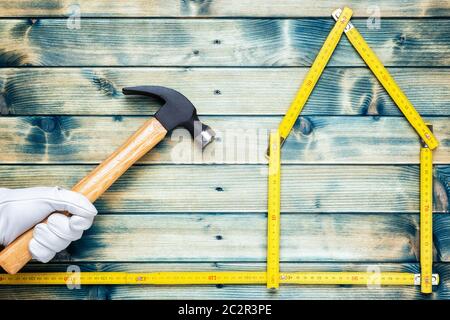 Blick von oben auf die eine Hand durch die Arbeit mit Leder Handschuhe geschützt, des Zimmermanns Hammer auf einem antiken Holztisch hält. Haus Konzept mit dem Messgerät. Ich Stockfoto