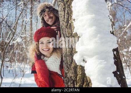 Verspieltes Paar versteckt sich hinter einem Baumstamm im Schnee und schaut in die Kamera Stockfoto
