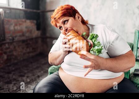 Fette Frau sitzt in einem Sessel und isst Sandwich, bulimic und Übergewicht. Ungesunde Lebensweise, Adipositas Stockfoto