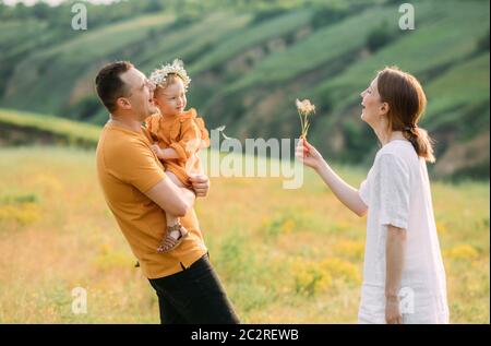 Junge Familie mit kleiner Tochter hat Spaß, bläst auf einen Löwenzahn und lacht fröhlich. Stockfoto