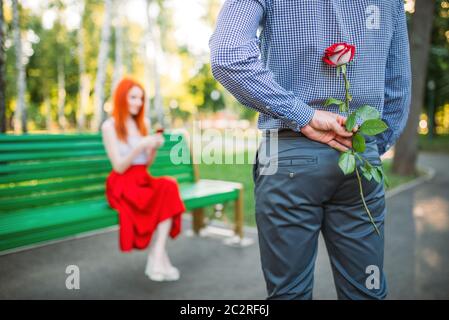 Attracrive Frau sitzt auf einer Bank im Park, Mann mit rose steht vor. Romantisches Date liebe paar Stockfoto