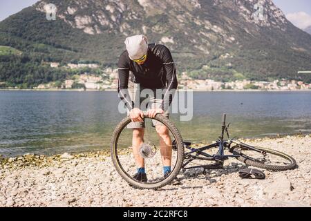 Fahrradreparatur. Mann Repariert Mountainbike. Radfahrer Mann in Schwierigkeiten Hinterrad Fall eines Unfalls. Mann repariert Fahrrad in der Nähe von See in Stockfoto