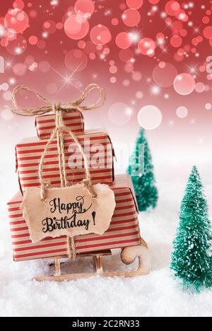 Schlitten mit Weihnachtsgeschenk und Label mit englischen Kalligraphie Happy Birthday. Weihnachtsbaum mit Schnee und rote Sekt Hintergrund Stockfoto