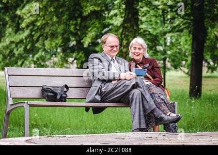 Glückliches positives älteres Paar lachend und Blick auf Handy auf Bank im Park an sonnigen Tag Stockfoto