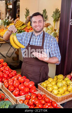 Lächelnder Mann bietet frische Bananen vor Kisten mit Tomaten und gelben Äpfeln. Lebensmittelgeschäft auf dem Hintergrund. Stockfoto