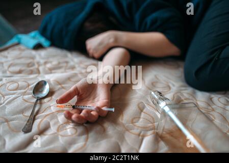 Weibliche Droggy mit Spritze in der Hand liegt im Bett, Flasche Alkohol und Löffel für die Dosis Vorbereitung auf Hintergrund. Drogenabhängigkeit Konzept, süchtig Menschen Stockfoto