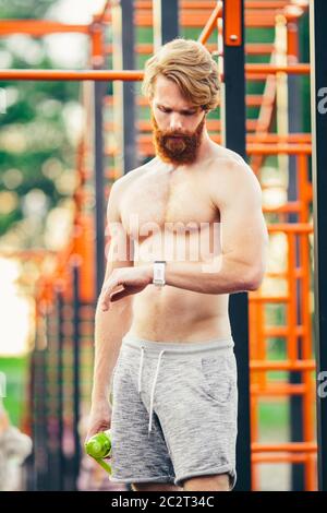 Sportmann, der die Uhr stehend Baumstäbchen Turnhalle. Junge Muskelsportler überprüfen Sportergebnisse auf Smart Watch nach einem guten Training Stockfoto