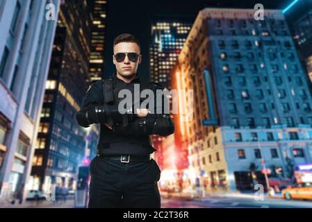 Polizeioffizier in Sonnenbrille und schwarzen Uniform mit Body Armor, Night city für den Hintergrund. Polizisten in spezielle Munition Stockfoto