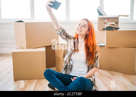 Lächelnde Frau sitzen auf dem Boden zwischen den Kartons und macht Selfie auf Handy-Kamera, Einweihungsparty. Umzug in neues Zuhause Stockfoto