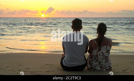 Silhouette eines hübschen jungen Paares sitzt am leeren Meeresstrand und beobachtet den Sonnenuntergang im tropischen Resort am Abend mit Blick auf die Rückseite Stockfoto