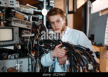 Seltsamer Ingenieur hält Bündel von Drähten mit verschiedenen Steckern im Labor. Elektrische Messgeräte im Hintergrund Laborausrüstung, Ingenieurwerkstatt Stockfoto