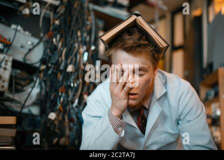 Müde seltsamer Ingenieur mit Buch auf dem Kopf schläft im Labor. Elektrische Messgeräte im Hintergrund Laborausrüstung, Ingenieurwerkstatt Stockfoto