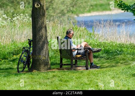 Männlicher Radfahrer sitzt, eine Pause auf der Bank, SMS am Telefon, Fahrrad von Baum gestützt - Scenic River Wharfe, Bolton Abbey Estate, Yorkshire England Großbritannien Stockfoto