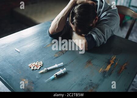 Männliche Junkie am Tisch mit Drogen und Spritze, grunge Inneneinrichtung im Hintergrund sitzen. Drogensucht Konzept, Leute süchtig Stockfoto