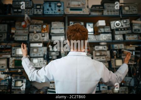 Seltsamer Wissenschaftler, der im Labor mit elektronischen Geräten im Regal steht, Rückansicht. Elektrische Messgeräte im Hintergrund Laborausrüstung, Ingenieurin Stockfoto