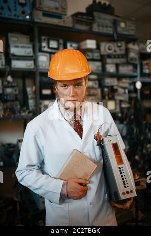 Seltsamer Wissenschaftler in einem Helm, der ein Buch und ein elektronisches Gerät hält, Ingenieur im Labor. Elektrische Messgeräte im Hintergrund Laborausstattung Stockfoto