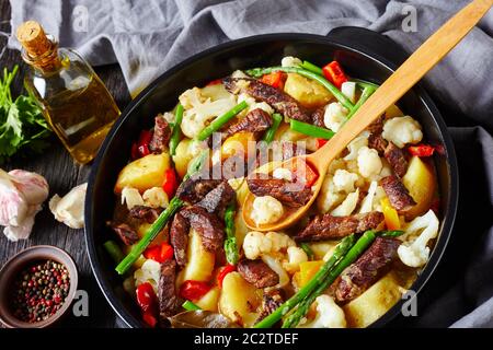 Rinderstreifen Eintopf mit neuen Kartoffeln, Spargel und Blumenkohl im schwarzen holländischen Ofen auf einem Holztisch, Landschaftsansicht, Nahaufnahme Stockfoto