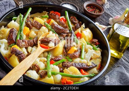 Würzige Rinderstreifen Eintopf mit neuen Kartoffeln, Spargel und Blumenkohl im schwarzen holländischen Ofen auf einem Holztisch, Landschaftsansicht, Nahaufnahme Stockfoto