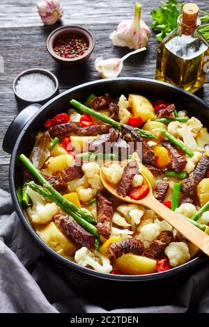 Rinderstreifen Eintopf mit neuen Kartoffeln, Spargel und Blumenkohl im schwarzen holländischen Ofen auf einem Holztisch, vertikale Ansicht, Nahaufnahme Stockfoto