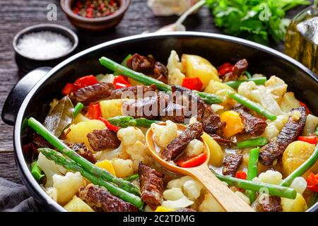 Nahaufnahme von würzigen Rinderstreifen Eintopf mit neuen Kartoffeln, Spargel und Blumenkohl in einem schwarzen holländischen Ofen auf einem Holztisch, Landschaftsansicht Stockfoto