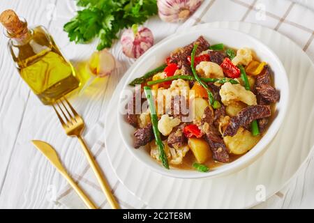 Sommer Rinderstreifen Eintopf mit neuen Kartoffeln, Spargel, Paprika-Stücke und Blumenkohl in einer weißen Schüssel auf einem Holztisch, amerikanische Küche, Landschaftsgärtnern Stockfoto