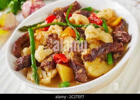 Nahaufnahme des Sommers Rinderstreifen Eintopf mit neuen Kartoffeln, Spargel und Blumenkohl in einer weißen Schüssel auf einem Holztisch, amerikanische Küche, Blick auf die Landschaft Stockfoto