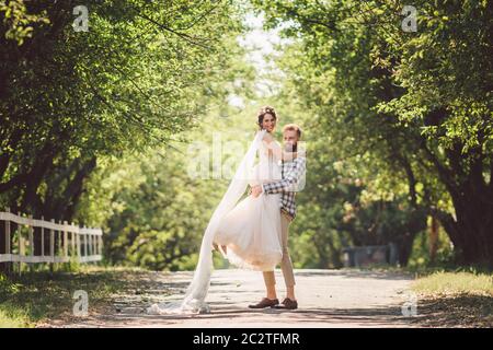 Glücklicher Bräutigam erhebt Braut im Sommerpark. Mann hob und umarmt Frau an ihren Armen. Frisch verheiratetes Paar im Park. Nur Heiratsfest Stockfoto