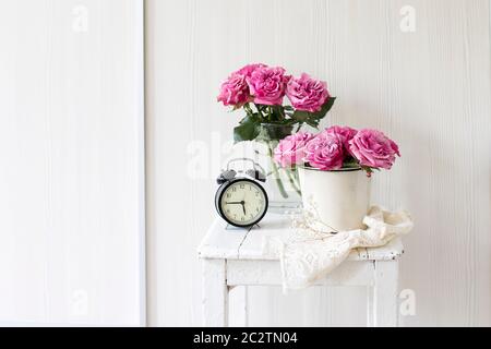 Ein Strauß rosa Rosen in einem Zinn weißen Eimer auf einem alten Hocker mit Wecker und einer Serviette mit Stickerei neben dem Bett auf einem beigen Hintergrund Stockfoto