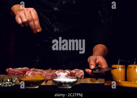 Der Küchenchef des Restaurants streut mit Currygewürzen und Basilikum auf schwarzem Hintergrund. Marinieren Schweinefleisch für Grill und Schinken. Vorderansicht. Stockfoto