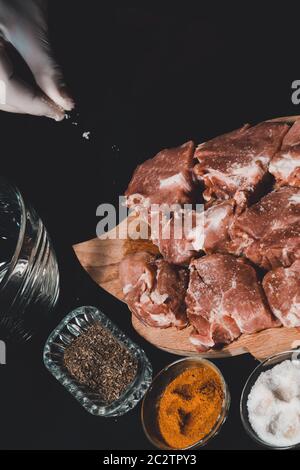 Der Küchenchef des Restaurants streut mit Currygewürzen und Basilikum auf schwarzem Hintergrund. Marinieren Schweinefleisch für Grill und Schinken. Draufsicht. Stockfoto