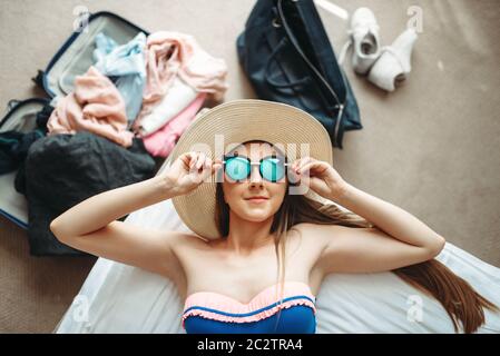 Junge Frau liegt im Badeanzug und Sonnenbrille, Ansicht von oben, eröffnete Koffer mit Kleidung für den Hintergrund. Die Gebühren für die Reise. Vorbereitung für den Sommer Vac Stockfoto