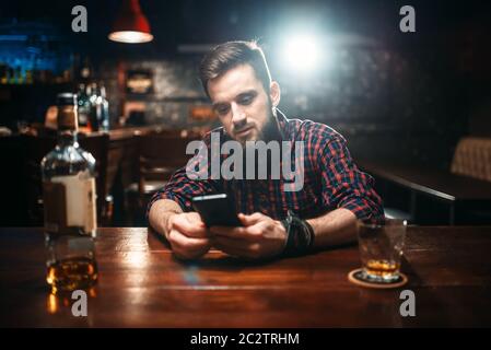 Lächelnd Mann an der Theke sitzen und über sein Handy, Entspannung mit Alkohol. Happy Guy Spaß, Freizeit in Pub Stockfoto