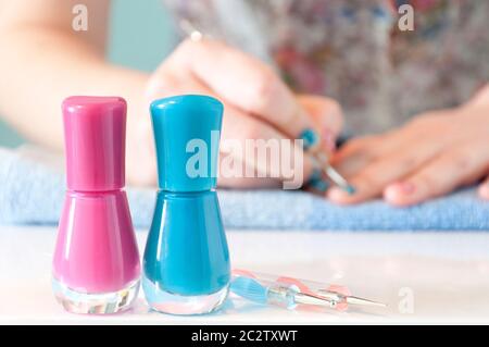 Im Vordergrund befinden sich zwei Nagellacke (ein rosa und ein blau) mit Maniküre-Werkzeugen. Im Hintergrund aus dem Fokus, Frauen Hände malen sie Stockfoto