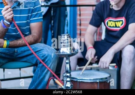 Nahaufnahme des Kopfes der Shisha aus Stahl. Ein Mann wird gesehen, wie er Shisha raucht, während ein anderer die Trommel spielt. Stockfoto