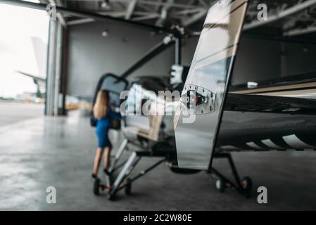 Weibliche Flight Attendant steht gegen Helikopter im Hangar mit geöffneten Toren. Stewardessen in Uniform in der Nähe von Copter. Private air transport Stockfoto