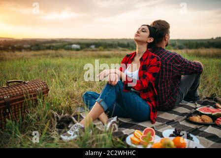 Der Mann und die Frau mit dem Rücken zueinander sitzen auf den Sonnenuntergang, Picknick im Feld. Romantischen Quark auf Sonnenuntergang, Paar im Freien Abendessen, glücklich relat Stockfoto