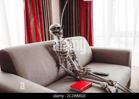 Lächelnde Skelett in Brille sitzt auf der Couch zwischen Buch und TV-Fernbedienung, Fenster- und rote Vorhänge für den Hintergrund. Lustiger Witz Stockfoto
