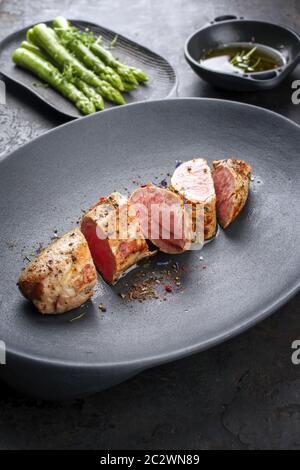 Gebratenes iberisches Schweinefilet in Scheiben geschnitten mit blanchierten grünen Spargel und Buttersauce mit Gewürzen als Nahaufnahme in einer gusseisernen Designplate Stockfoto