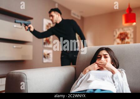 Verängstigte Frau versteckt sich auf der Couch gegen Mörder in schwarzer Kleidung mit der Waffe in der Hand. Gangster die Wohnung eingedrungen. Raub zu Hause Stockfoto