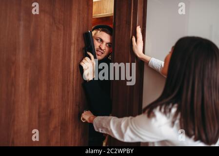 Verängstigte Frau, die versucht, die Tür zu schließen, Mörder in schwarzer Kleidung mit der Waffe in der Hand die Wohnung einzudringen. Raub zu Hause Stockfoto